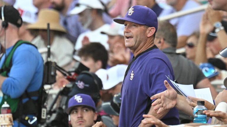 An LSU baseball coach stands looking ahead on the field as team members and fans discuss the transfer portal in the stadium.