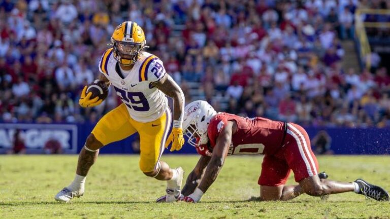 An LSU player in yellow and purple runs with the ball as an Arkansas defender in red tries to tackle him.