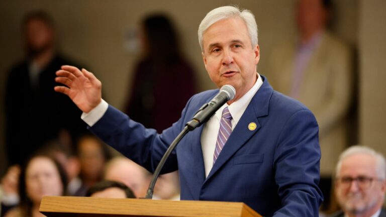 The LSU President gestures while speaking at a podium with a microphone; an audience sits in the background.