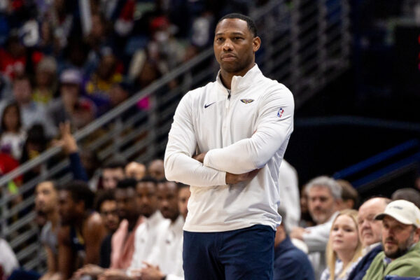 A basketball coach in a white zip-up jacket stands courtside with arms crossed as spectators watch the game.