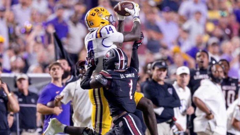 An LSU football player in yellow uniform leaps to catch a pass above a South Carolina defender during a game.