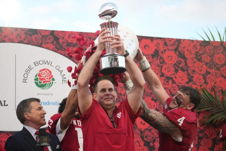 An Indiana coach lifts a trophy as players in red uniforms celebrate beside him during the Rose Bowl Game.