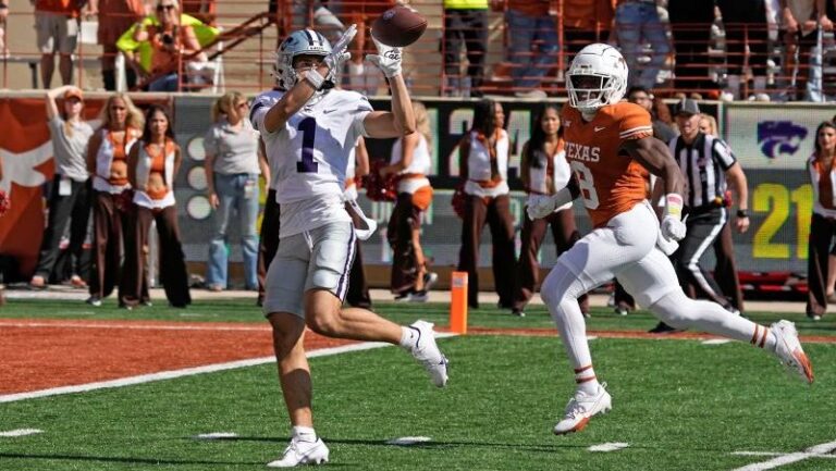 Kansas State’s Jayce Brown catches a pass in a white and purple uniform as an opposing player in orange runs toward him on the field.