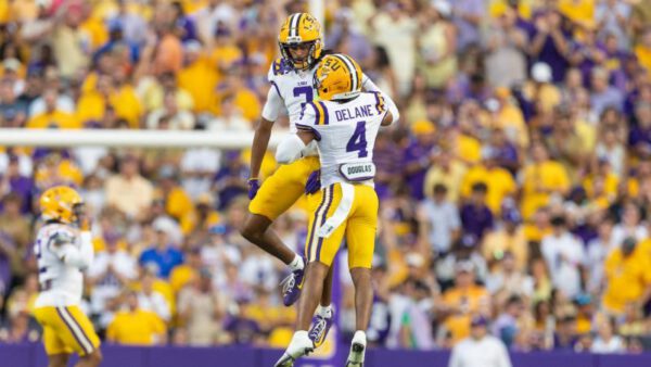 Two LSU football players in uniform jump and celebrate together on the field as fans watch from the stands in the background.