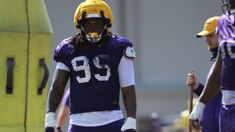 LSU football player in a yellow helmet and purple #99 jersey stands on the field with teammates during practice.