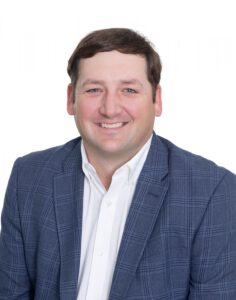 Smiling man with short brown hair in a white shirt and blue plaid blazer, standing against a plain white background.