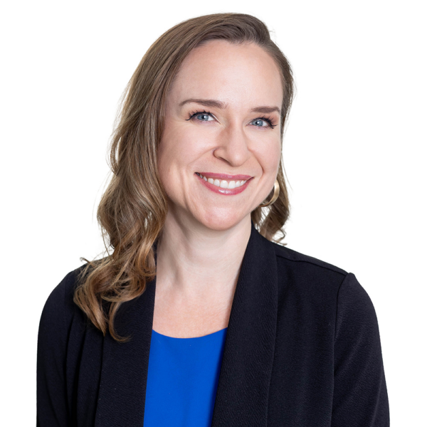 Smiling woman with long light brown hair, wearing a blue top and black blazer, stands before a plain white background.