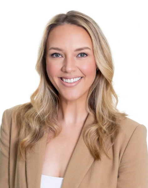 A woman with long blonde hair smiles in a tan blazer and white top against a simple white background.