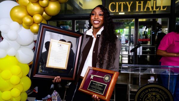 A woman stands by Baton Rouge City Hall, holding two framed awards near gold and white balloons and a nearby podium.