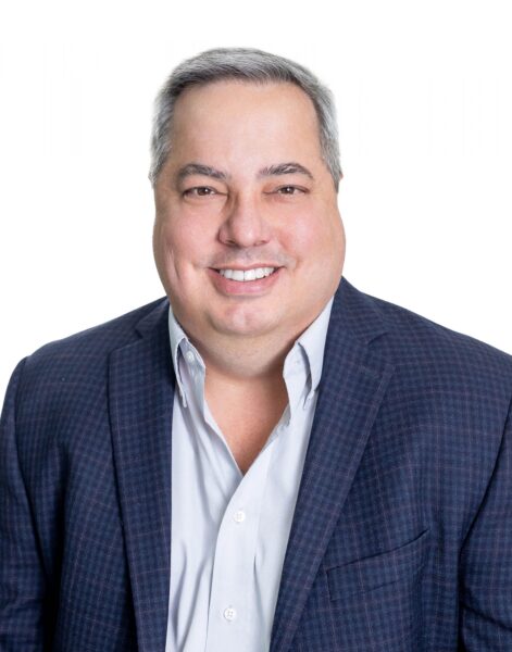 Charles Hanagriff, with short gray hair in a dark checkered blazer and light shirt, smiles at the camera against a white background.