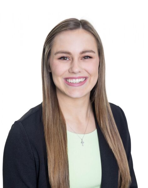 A woman with long brown hair smiles at the camera, dressed in a black blazer and light green top, standing before a white background.