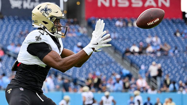 A Saints player extends his arms to catch a football at OTA day 4, with teammates and empty stadium seats in the background.
