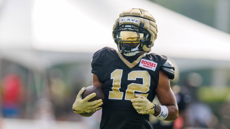 Number 12 in black and gold gear holds a football during practice on a sunny day.