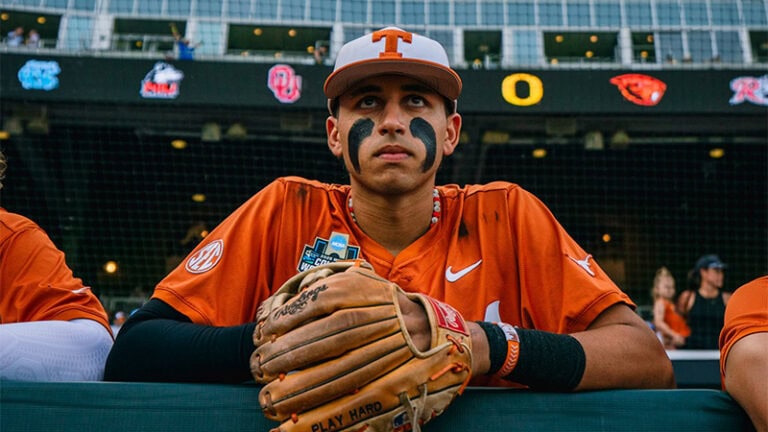 A Texas baseball player in an orange uniform and cap sits in the dugout holding a glove, with stadium seats visible behind him.