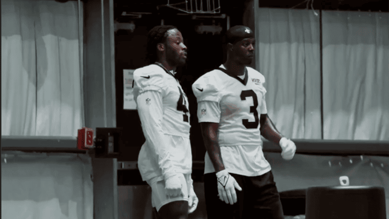 Two football players in white uniforms, both wearing gloves, stand indoors at Saints OTA looking in the same direction.