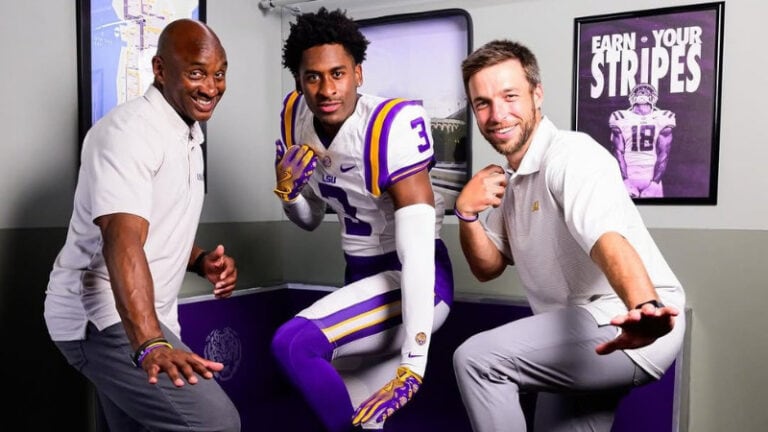 Three men pose playfully indoors; one is in an LSU football uniform, while the other two wear light polo shirts and pants.