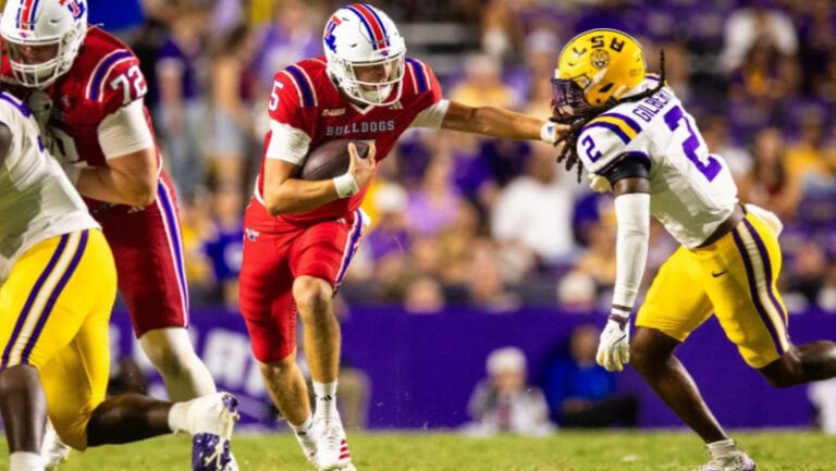 A Louisiana Tech player in red runs with the ball, pushing away a yellow and purple defender during a football game.