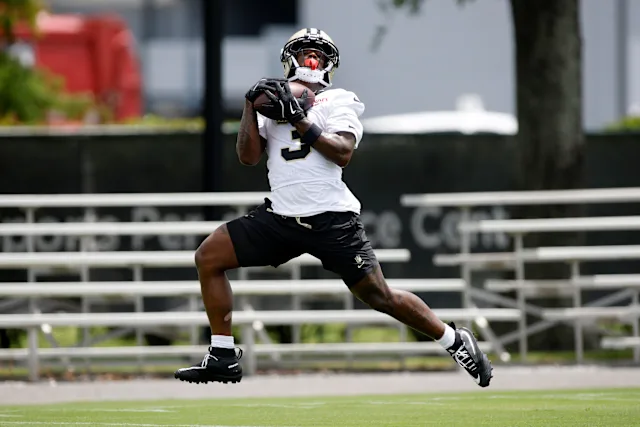 Travis Etienne Jr., wearing a white jersey and black shorts, catches a football during practice on a field with empty bleachers behind him.