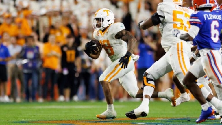 A Tennessee player in a white and orange uniform runs with the ball as blue-uniformed opponents chase him during a football game.