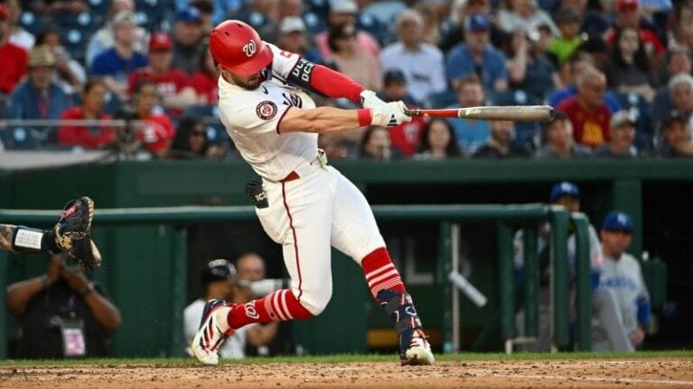 A baseball player in a white and red uniform swings his bat during an MLB game as fans look on from the stands.