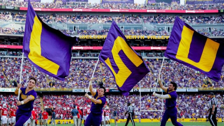 Tre Segarra and two others wave large purple and gold "LSU" flags on a football field before a full stadium crowd.