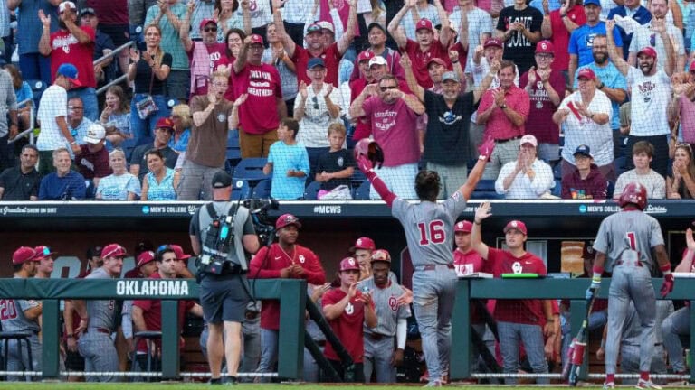 A baseball player in a gray uniform, number 16, raises his arms to celebrate with cheering fans after an SEC win in Omaha.