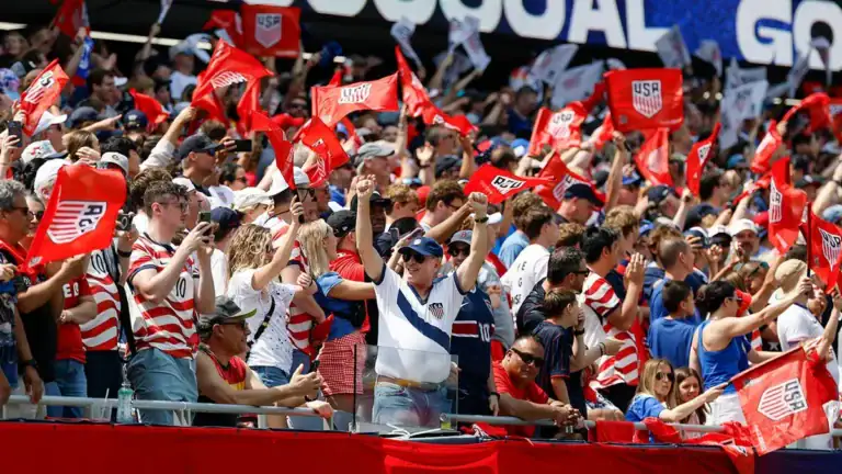 Fans in red, white, and blue cheer and wave USA flags at a World Cup stadium during the day.