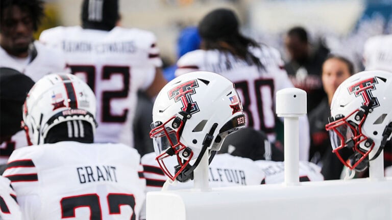 Texas Tech football helmets sit on a rack as players in white uniforms gather nearby on the sideline.