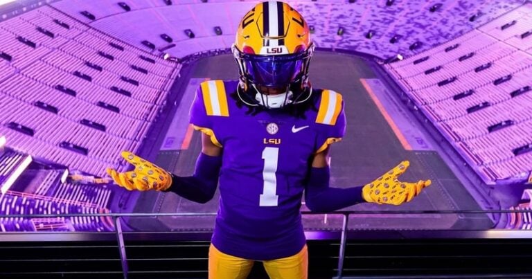 An LSU football player stands with arms outstretched in an empty stadium illuminated by purple lights.