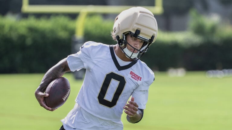 A football player in a white number 0 jersey runs with the ball at Saints Training Camp, helmet on, across a grassy field.
