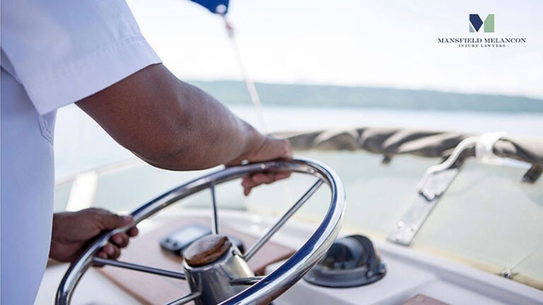 A person in a white shirt steers a boat with a wooden wheel as water and shoreline stretch out in the background.