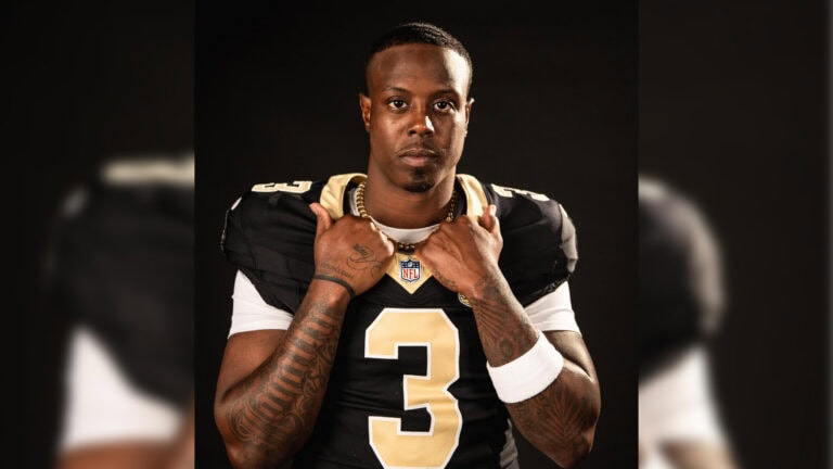 A football player, similar to Travis Etienne Jr., holds his collar and looks confidently at the camera.