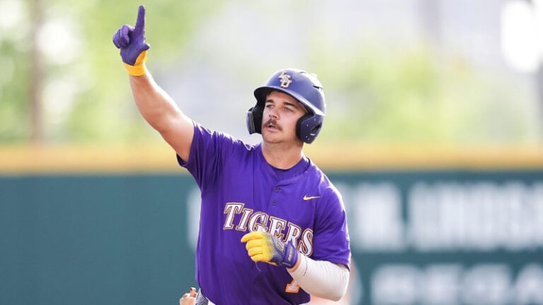 Baseball player in a purple Tigers uniform, wearing helmet and gloves, points upward on the field.