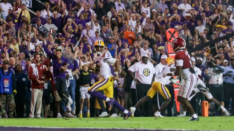 A football player in white and yellow runs with the ball at Tiger Stadium, while fans cheer in the stands behind him.