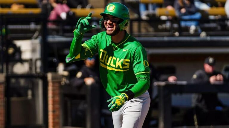 A baseball player in a green Oregon Ducks uniform gestures during a game, with spectators watching in the background.