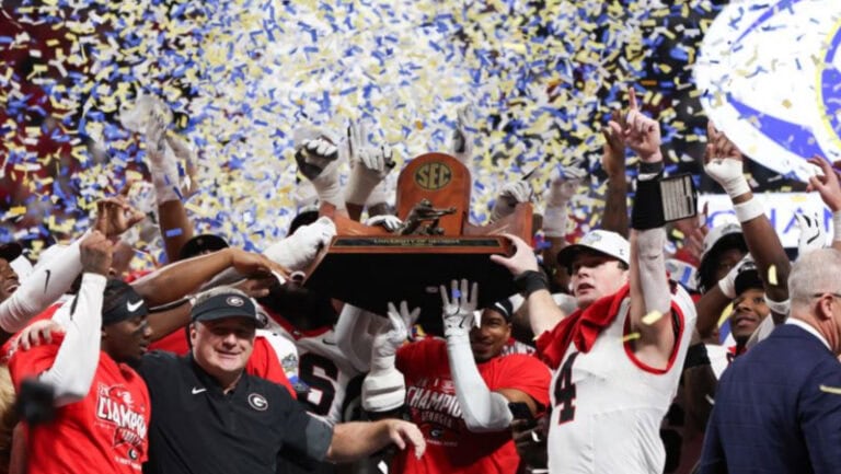 Players in red and white uniforms celebrate a conference title win, lifting the SEC trophy as blue and yellow confetti falls around them.