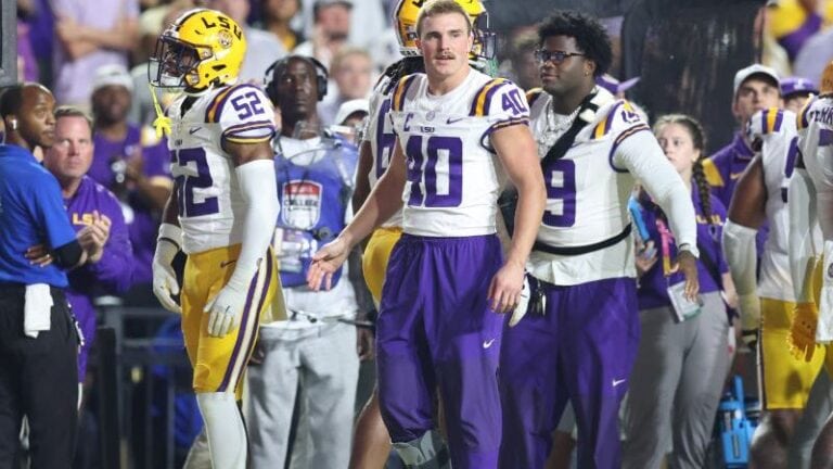 LSU football players in uniform watch from the sideline during a game, focused on the field after Weeks’ injury.