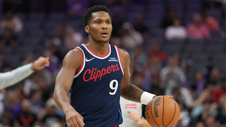 A Clippers guard dribbles a Wilson basketball on court, with a blurred crowd and Pelicans fans visible in the background.