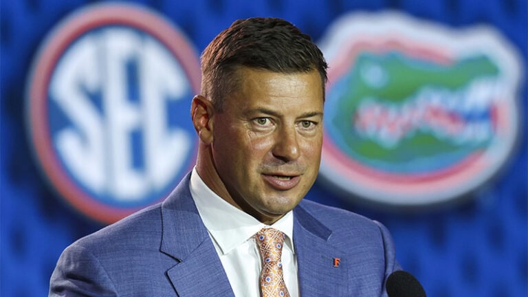 Jon Sumrall speaks at a podium, with blurred SEC and Florida Gators logos in the background.