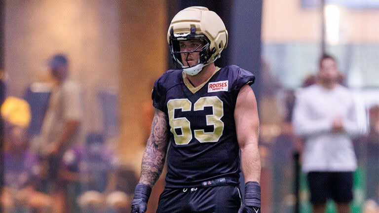 A football player in a black and gold number 63 uniform stands on an indoor field during practice.