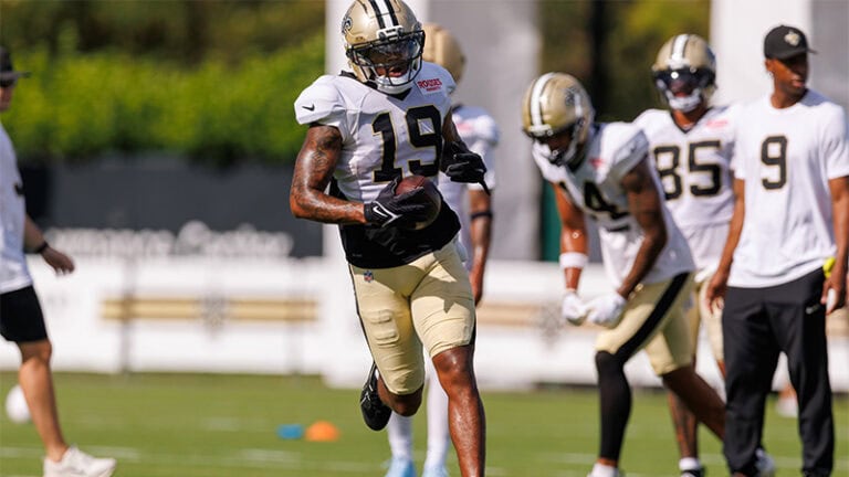 A Saints player in white and gold runs with the ball at training camp while teammates and coaches observe in the background.