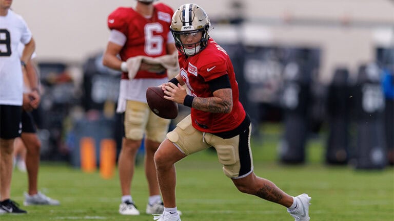 A football player in a red jersey runs with the ball at NFL preseason practice, teammates on the grassy field behind him.