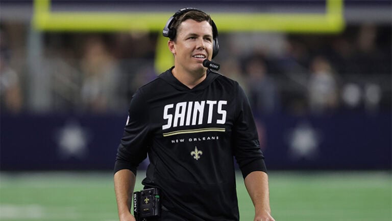 A football coach in a headset and Saints sweatshirt stands on the field, with blurred stadium seats in the background.