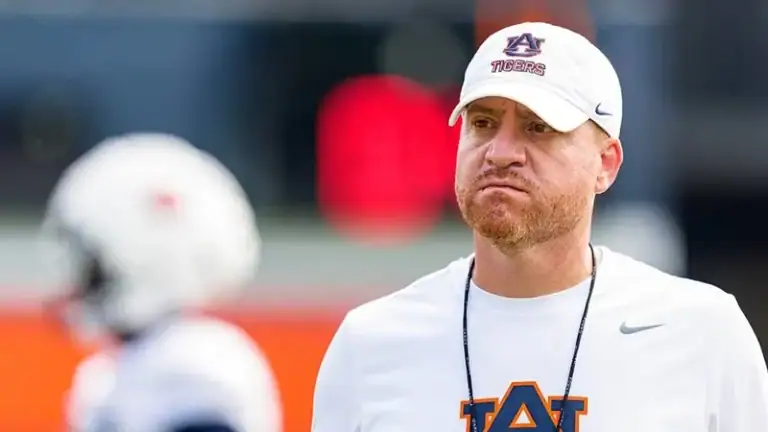 Alex Golesh stands outdoors, watching SEC fall camp as players prepare and gear up in the background.