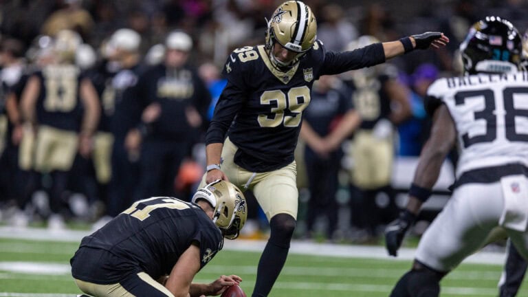 A Saints player in black lines up for a field goal as his teammate holds the ball during a preseason game.