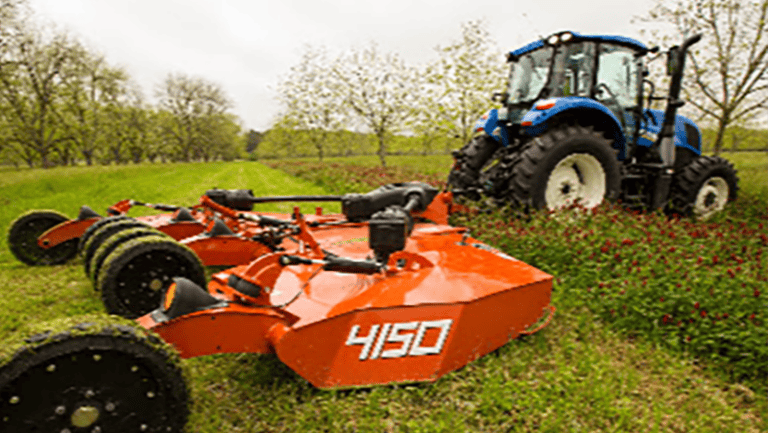 A blue tractor pulls an orange mower labeled 4150, mowing a green field for deer season with trees in the background.