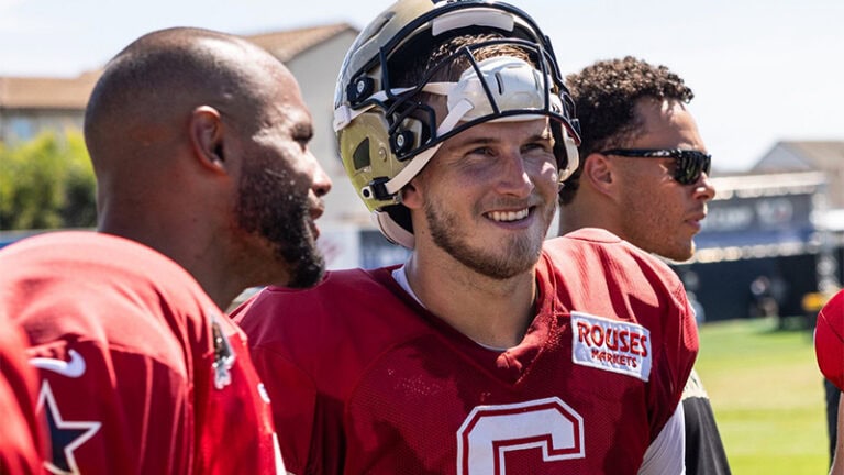 Three football players in red jerseys stand outdoors, one wearing a helmet and smiling, with houses and blue sky behind them.