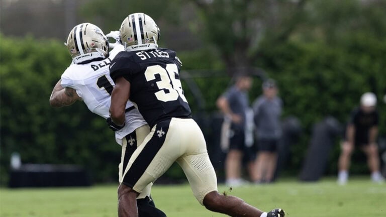 Two Saints players in padded uniforms practice on a grassy field as blurred spectators watch in the background.