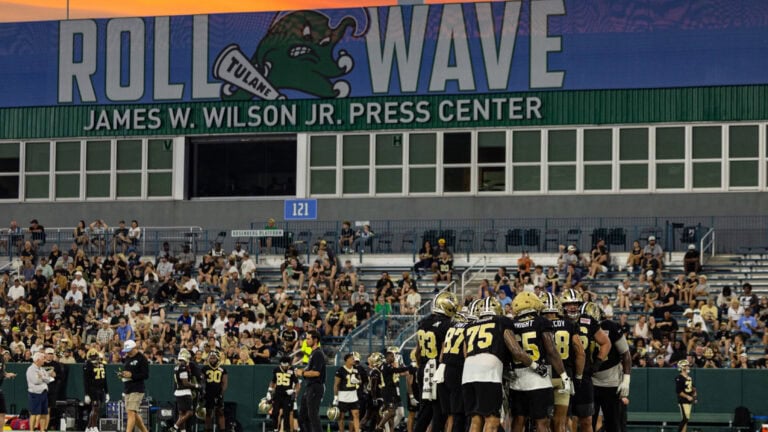 Saints players huddle at training camp, with a “Roll Wave” Tulane sign and excitement over the kicker battle in the background.