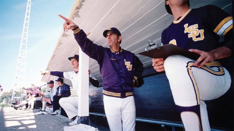 A baseball coach in an LSU jacket stands in the dugout, pointing, as players watch him with focus, reflecting Skip Bertman’s legacy.